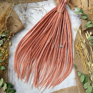 Puede incluir: Un conjunto de rastas sintéticas de color rosa y blanco degradado. Las rastas están dispuestas en forma de abanico y tienen un degradado de rosa claro a blanco.