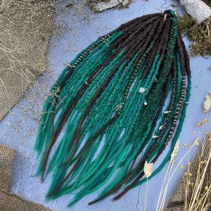 Puede incluir: Conjunto de rastas hechas a mano en tonos marrón y verde turquesa. Las rastas están decoradas con abalorios y cuentas plateadas. Los extremos de las rastas son de un vibrante color verde turquesa. Presentadas sobre una superficie azul claro.