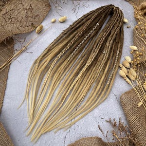 Puede incluir: Un conjunto de rastas largas sintéticas en un color degradado de marrón claro a rubio. Las rastas están trenzadas y tienen cuentas doradas.