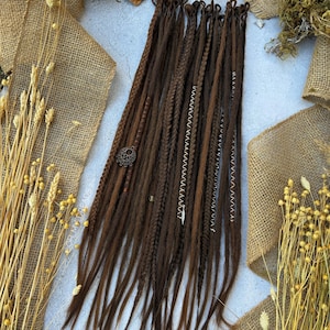 Puede incluir: Un conjunto de rastas sintéticas marrones con varios adornos, incluyendo cuentas y detalles metálicos. Las rastas están dispuestas en una fila sobre una superficie de color claro.