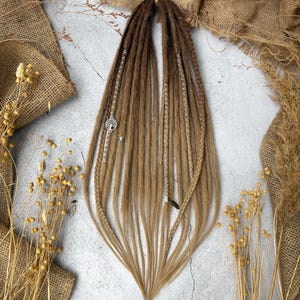 Puede incluir: Dos conjuntos de rastas marrones y rubias con detalles plateados y abalorios decorativos. Las rastas están dispuestas sobre un fondo neutro con tela de yute y plantas secas. Las extensiones de cabello son una mezcla de marrón y rubio.