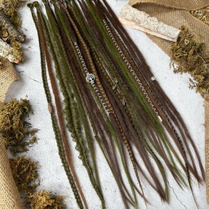 May include: A collection of handmade dreadlocks in shades of brown and green. Some dreads are adorned with silver-colored beads and a tree of life charm. The dreadlocks are arranged on a light-colored surface with burlap and moss.