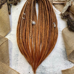 Puede incluir: Un conjunto de rastas sintéticas marrones con cuentas doradas y plateadas. Las rastas están dispuestas en forma de abanico y están sobre una superficie blanca.