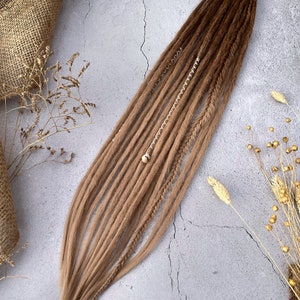 Puede incluir: Un conjunto de rastas sintéticas largas de color marrón con cuentas y una sección trenzada. Las rastas están dispuestas sobre una superficie gris claro.