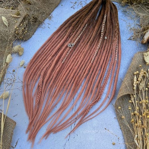 Puede incluir: Un conjunto de rastas largas sintéticas con un esquema de color degradado de dos tonos. Las rastas son marrones en la parte superior y se desvanecen a un color rosa claro en la parte inferior. Las rastas están decoradas con cuentas plateadas y un dije plateado.