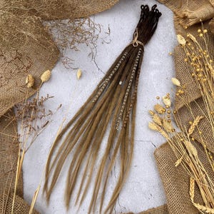 Puede incluir: Un conjunto de rastas sintéticas marrones con cuentas doradas. Las rastas están atadas juntas con una cuerda marrón.