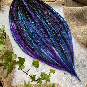 Puede incluir: Un conjunto de rastas sintéticas en tonos negro, azul y morado. Las rastas están trenzadas y tienen cuentas plateadas.