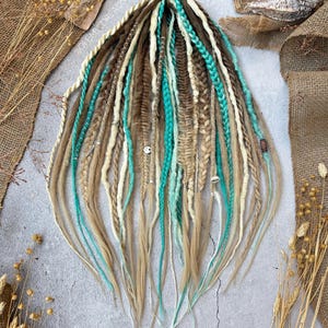 Puede incluir: Conjunto de rastas hechas a mano en tonos marrón, rubio y turquesa. Algunas están trenzadas, otras torcidas, y algunas adornadas con pequeñas cuentas y abalorios. Las rastas están dispuestas sobre una superficie neutra con arpillera y plantas secas.