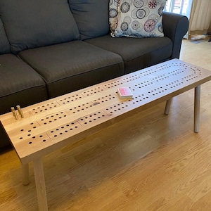 May include: A wooden cribbage board game table with a natural wood finish. The table has a rectangular top with a cribbage board design and four legs. There are two sets of pegs on the table, and a deck of cards is sitting on the table.