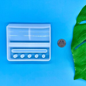 May include: White silicone mold for resin crafts. The mold has a rectangular shape with two compartments and five round holes.