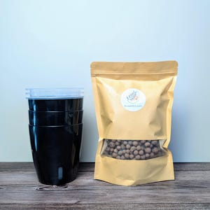 May include: A brown paper bag with a clear window showing brown clay pebbles. The bag has a white logo with the text "seaddict.com". Three black plastic pots are stacked on top of each other.