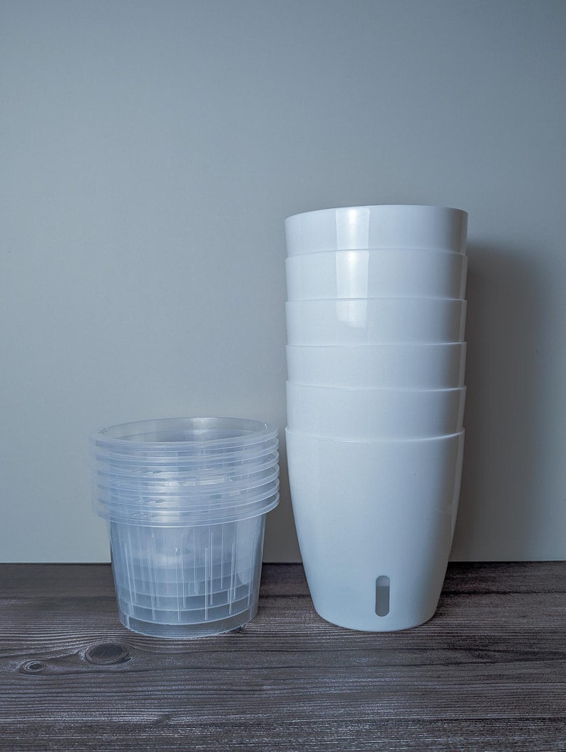 a stack of 6 white self watering pots. White outer pot and clear inner pot sit next to each other.