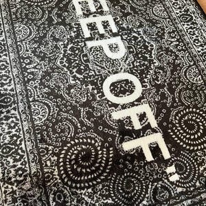 K&ouml;nnte beinhalten: Ein rechteckiger Teppich mit einem schwarz-wei&szlig;en Paisley-Muster und dem Text KEEP OFF, der in gro&szlig;en wei&szlig;en Gro&szlig;buchstaben in der Mitte aufgedruckt ist. Das Design umfasst komplexe wirbelnde Motive und eine dekorative Bord&uuml;re an den R&auml;ndern.