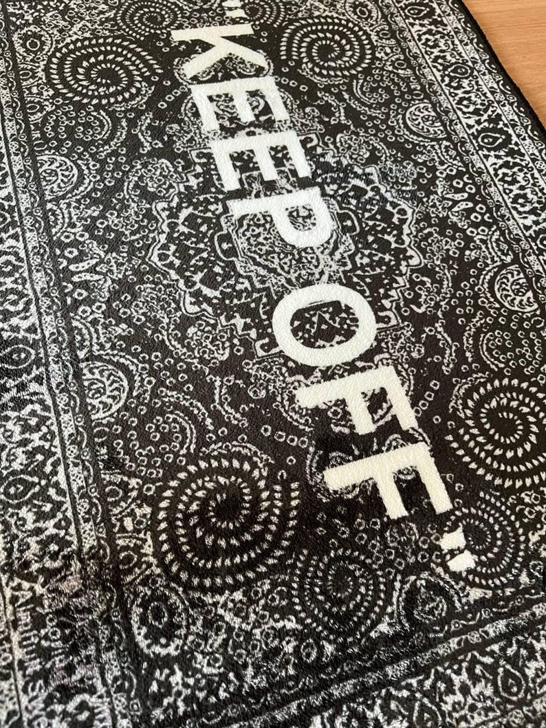 K&ouml;nnte beinhalten: Ein rechteckiger Teppich mit einem schwarz-wei&szlig;en Paisley-Muster und dem Text KEEP OFF, der in gro&szlig;en wei&szlig;en Gro&szlig;buchstaben in der Mitte aufgedruckt ist. Das Design umfasst komplexe wirbelnde Motive und eine dekorative Bord&uuml;re an den R&auml;ndern.