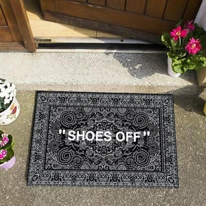 K&ouml;nnte beinhalten: Schwarz-wei&szlig;er Fu&szlig;abtreter mit einem komplizierten Muster und dem Text "SHOES OFF" in Wei&szlig;.