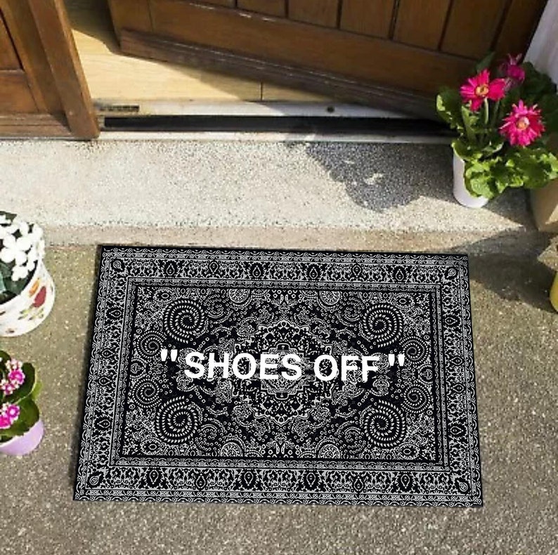 K&ouml;nnte beinhalten: Schwarz-wei&szlig;er Fu&szlig;abtreter mit einem komplizierten Muster und dem Text "SHOES OFF" in Wei&szlig;.
