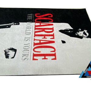 May include: A black and white rug with a picture of Al Pacino as Tony Montana from the movie Scarface. The text "Scarface" is in red and the text "The World Is Yours" is in black.