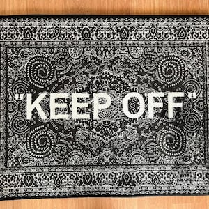 K&ouml;nnte beinhalten: Ein rechteckiger schwarz-wei&szlig;er Teppich mit einem detaillierten Paisley-Muster auf der gesamten Oberfl&auml;che. Der Text "KEEP OFF" ist in gro&szlig;en, fetten, wei&szlig;en Gro&szlig;buchstaben in der Mitte des Teppichs gedruckt und von einem dekorativen Rand umgeben.