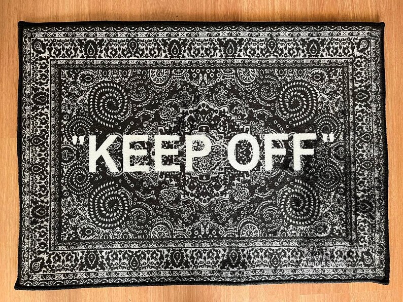 K&ouml;nnte beinhalten: Ein rechteckiger schwarz-wei&szlig;er Teppich mit einem detaillierten Paisley-Muster auf der gesamten Oberfl&auml;che. Der Text "KEEP OFF" ist in gro&szlig;en, fetten, wei&szlig;en Gro&szlig;buchstaben in der Mitte des Teppichs gedruckt und von einem dekorativen Rand umgeben.