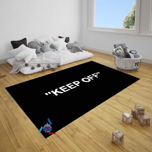 May include: Black and white rug with the text "KEEP OFF" in white letters. The rug is on a wooden floor in a room with a white wall and a window.