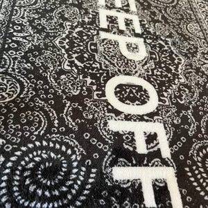 K&ouml;nnte beinhalten: Ein schwarz-wei&szlig;er rechteckiger Teppich mit einem detaillierten Paisley-Muster. Der Text "KEEP OFF" ist in gro&szlig;en, fetten wei&szlig;en Gro&szlig;buchstaben vertikal &uuml;ber die Mitte des Teppichs gedruckt und bildet einen Kontrast zum dunklen Hintergrund.