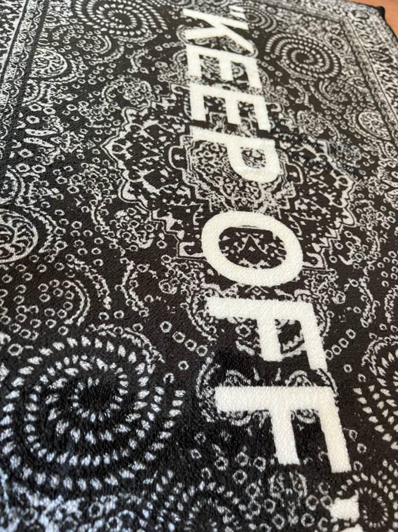 K&ouml;nnte beinhalten: Ein schwarz-wei&szlig;er rechteckiger Teppich mit einem detaillierten Paisley-Muster. Der Text "KEEP OFF" ist in gro&szlig;en, fetten wei&szlig;en Gro&szlig;buchstaben vertikal &uuml;ber die Mitte des Teppichs gedruckt und bildet einen Kontrast zum dunklen Hintergrund.