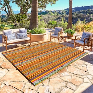 May include: A colorful outdoor rug with a geometric pattern in shades of orange, yellow, red, blue, and green. The rug is laid out on a patio with two wooden chairs and a table.