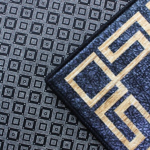 Może przedstawiać: Zbliżenie na dwa dywany o r&oacute;żnych wzorach. G&oacute;rny dywan ma czarne tło z białym geometrycznym wzorem. Dolny dywan ma ciemnoniebieskie tło z złotym geometrycznym wzorem. Słowo "SAMPLE" jest odciśnięte na czerwono na dolnym dywanie.