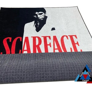 May include: A black and white rug with a red "Scarface" title and a portrait of Al Pacino as Tony Montana. The rug has a gray backing.