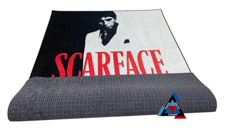 May include: A black and white rug with a red "Scarface" title and a portrait of Al Pacino as Tony Montana. The rug has a gray backing.