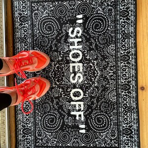 K&ouml;nnte beinhalten: Eine schwarz-wei&szlig;e gemusterte Fu&szlig;matte mit dem Text "Shoes Off!" in Wei&szlig;. Zwei rot-schwarze Schuhe befinden sich auf der Fu&szlig;matte.