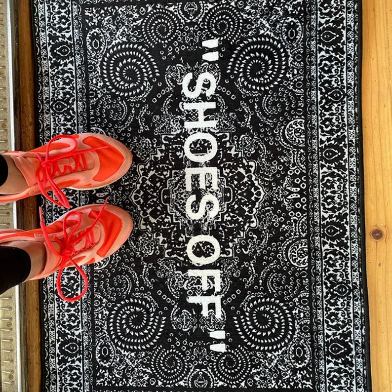 K&ouml;nnte beinhalten: Eine schwarz-wei&szlig;e gemusterte Fu&szlig;matte mit dem Text "Shoes Off!" in Wei&szlig;. Zwei rot-schwarze Schuhe befinden sich auf der Fu&szlig;matte.