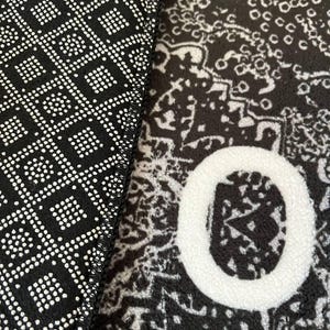 K&ouml;nnte beinhalten: Ein schwarz-wei&szlig;er Teppich mit einer strukturierten Oberfl&auml;che, einem sich wiederholenden komplizierten Muster und einem gro&szlig;en wei&szlig;en Buchstaben O. Die Unterseite des Teppichs ist schwarz mit einem geometrischen Gittermuster aus kleinen Quadraten und Punkten.