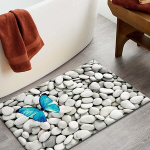 May include: A white bath mat with a blue butterfly and a white pebble design. The mat is placed on a wooden floor next to a white bathtub.