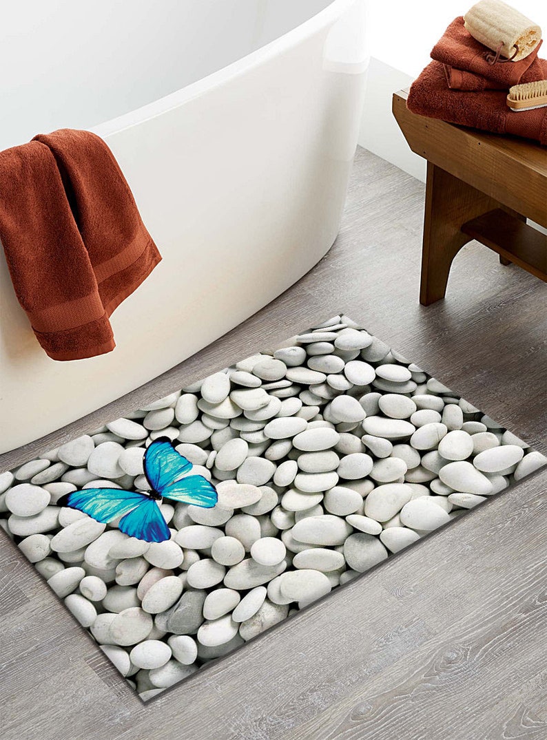 May include: A white bath mat with a blue butterfly and a white pebble design. The mat is placed on a wooden floor next to a white bathtub.