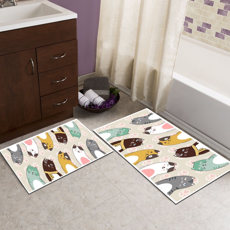 May include: Two bath mats with a beige background and a pattern of cartoon cats in various colours, including brown, yellow, green, and grey. The cats are upside down with their paws in the air. The mats have a light brown border.