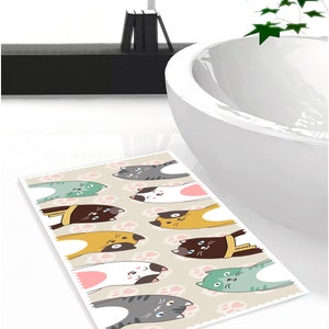 May include: A beige bath mat with a pattern of pink paw prints and nine cartoon cats in various colours and poses. The cats are looking up at the viewer.