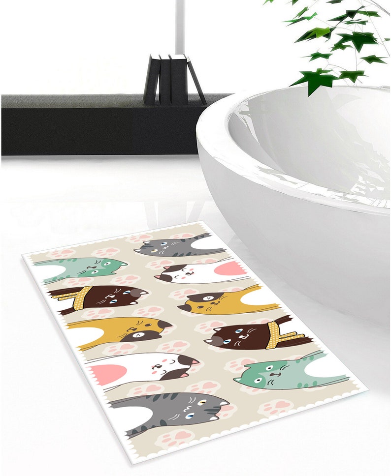 May include: A beige bath mat with a pattern of pink paw prints and nine cartoon cats in various colours and poses. The cats are looking up at the viewer.