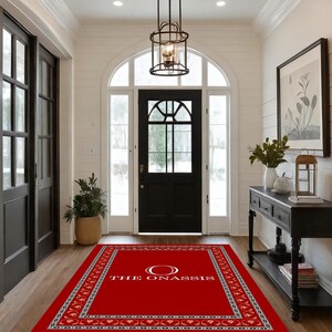 May include: A red rectangular welcome mat with a decorative border and the text "THE ONASSIS" in white. The mat is placed in a bright entryway with a black door and a decorative light fixture.