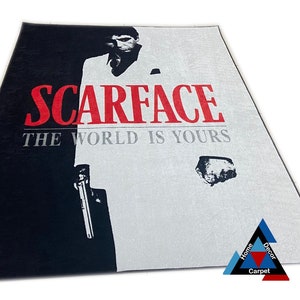 May include: Black and white rug with a graphic of a man in a white suit holding a gun. The text "Scarface" and "The World Is Yours" is printed in red on the rug.