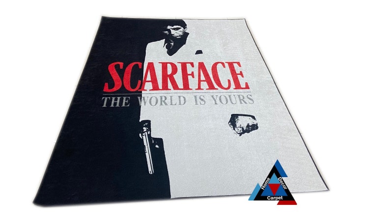 May include: Black and white rug with a graphic of a man in a white suit holding a gun. The text "Scarface" and "The World Is Yours" is printed in red on the rug.