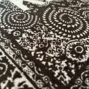 K&ouml;nnte beinhalten: Ein schwarz-wei&szlig;es Textil mit einem sich wiederholenden Paisley-Muster aus komplexen Wirbeln und floralen Motiven. Der Stoff hat eine weiche, strukturierte Oberfl&auml;che und zeigt den teilweisen wei&szlig;en Schriftzug "KEEP" in serifenloser Schrift.
