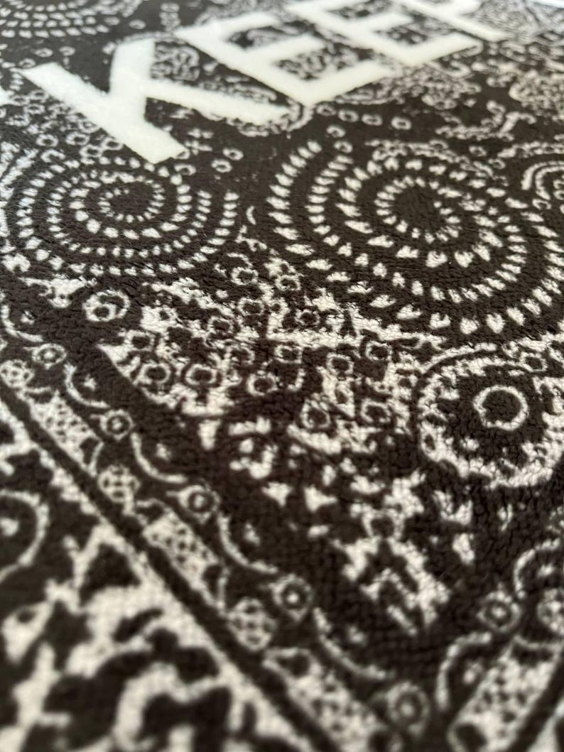 K&ouml;nnte beinhalten: Ein schwarz-wei&szlig;es Textil mit einem sich wiederholenden Paisley-Muster aus komplexen Wirbeln und floralen Motiven. Der Stoff hat eine weiche, strukturierte Oberfl&auml;che und zeigt den teilweisen wei&szlig;en Schriftzug "KEEP" in serifenloser Schrift.