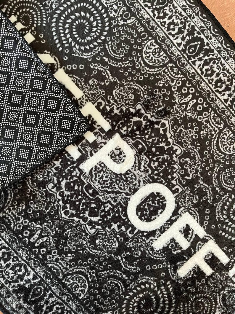 K&ouml;nnte beinhalten: Ein schwarz-wei&szlig;er Teppich mit einem detaillierten Paisley-Muster und dem Text "RIP OFF" in fetten wei&szlig;en Gro&szlig;buchstaben. Die Unterseite des Teppichs ist schwarz und weist ein strukturiertes, rutschfestes Gittermuster aus kleinen Quadraten und Punkten auf.