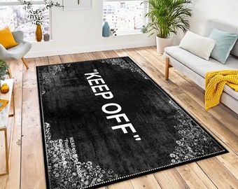 May include: A rectangular black rug with the words "KEEP OFF" in large white letters. The rug has a distressed texture and a floral border. The rug is placed on a wooden floor in a living room setting.