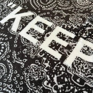 K&ouml;nnte beinhalten: Ein schwarz-wei&szlig;er Stoff mit einem traditionellen Paisley-Muster aus komplexen Wirbeln und floralen Motiven. Das Wort KEEP ist in gro&szlig;en, fetten, wei&szlig;en Gro&szlig;buchstaben in der Mitte der strukturierten Materialoberfl&auml;che aufgedruckt.