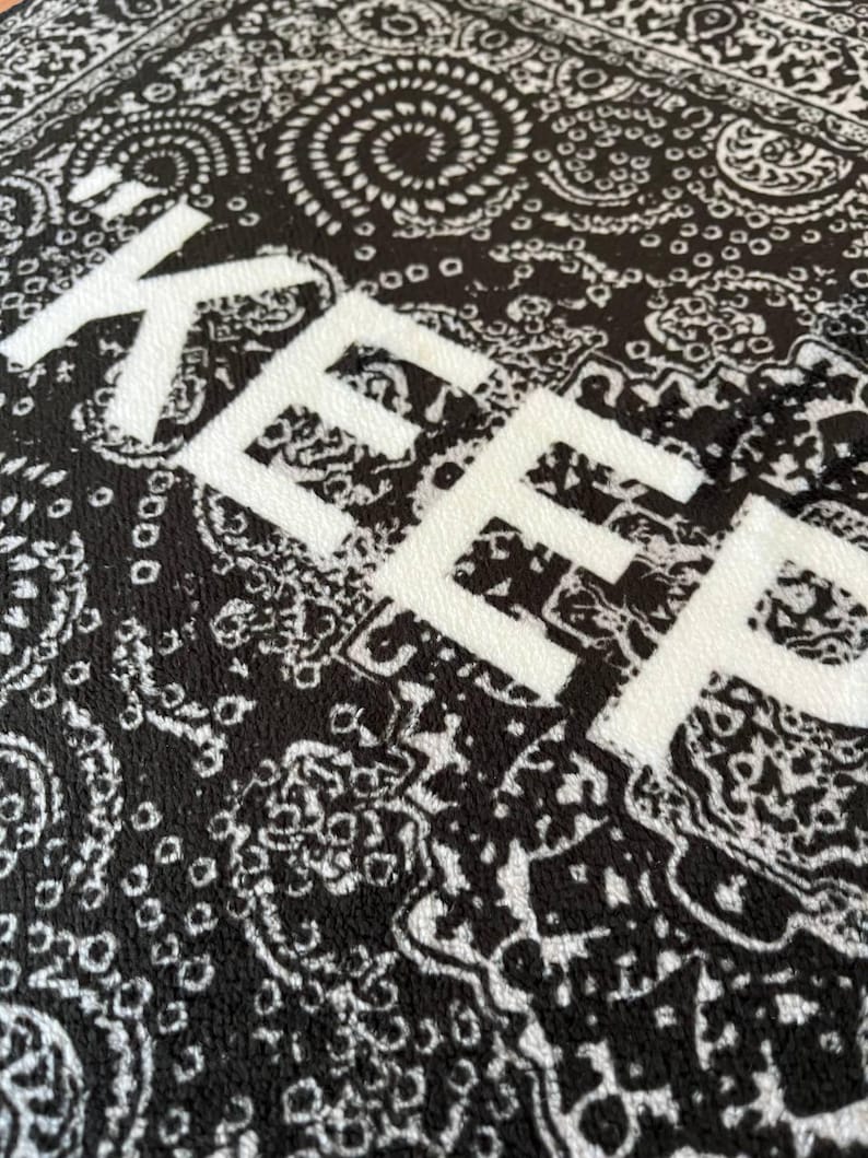 K&ouml;nnte beinhalten: Ein schwarz-wei&szlig;er Stoff mit einem traditionellen Paisley-Muster aus komplexen Wirbeln und floralen Motiven. Das Wort KEEP ist in gro&szlig;en, fetten, wei&szlig;en Gro&szlig;buchstaben in der Mitte der strukturierten Materialoberfl&auml;che aufgedruckt.