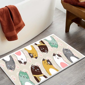 May include: A beige bath mat with a white border features a repeating pattern of pink paw prints and cartoon cats in various colours, including yellow, brown, grey, white, and green. The cats are all facing upwards with their paws in the air.