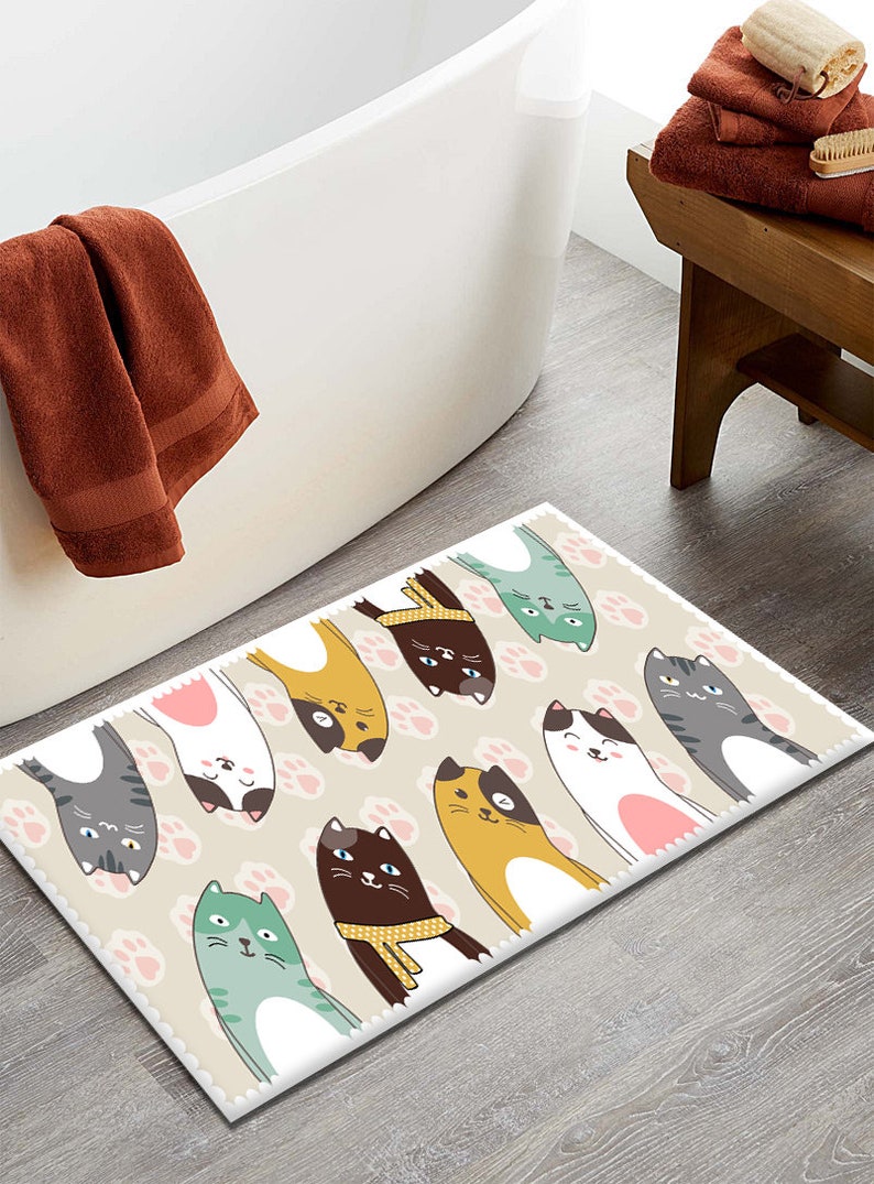 May include: A beige bath mat with a white border features a repeating pattern of pink paw prints and cartoon cats in various colours, including yellow, brown, grey, white, and green. The cats are all facing upwards with their paws in the air.