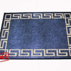 Puede incluir: Una alfombra azul con un borde de clave griega beige. La alfombra tiene una superficie texturizada.